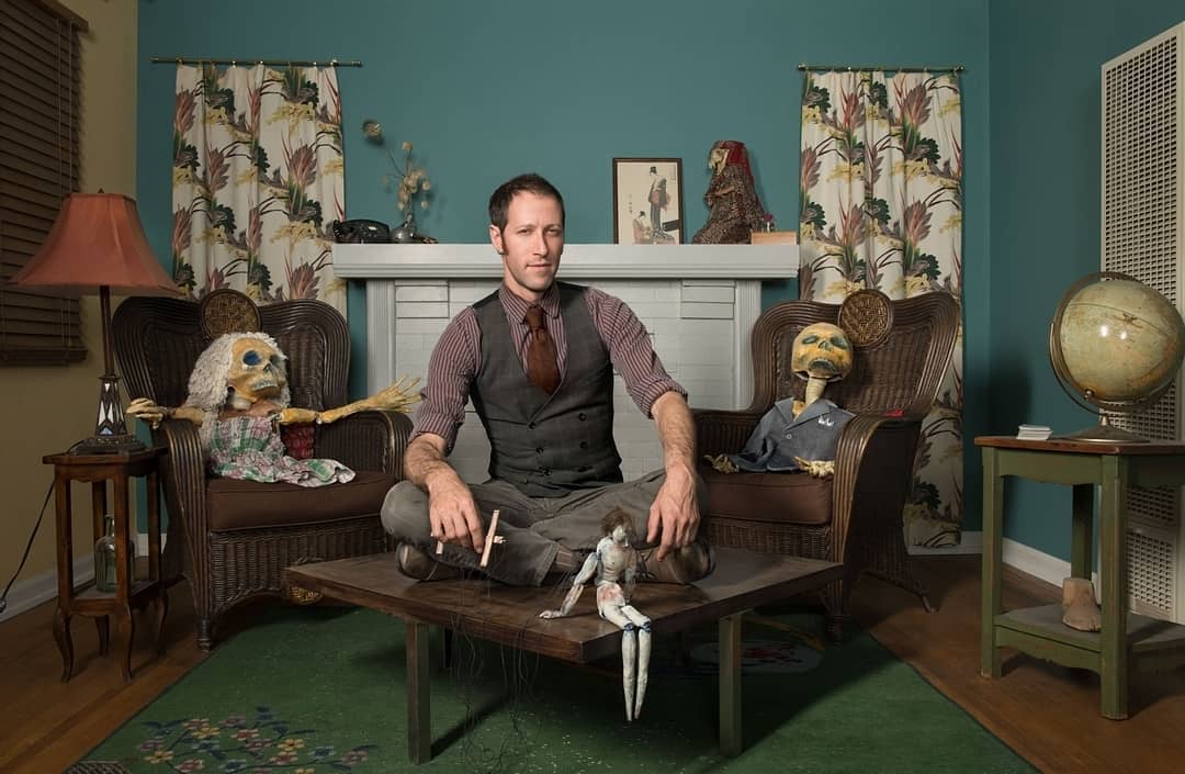 Man sits with puppets in an eclectic living room setting with teal walls, floral curtains, and vintage props in eye-level shot.